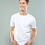 Portrait of happy young man wearing white t-shirt, standing with hands in pockets and smiling. Studio shot, grey background.